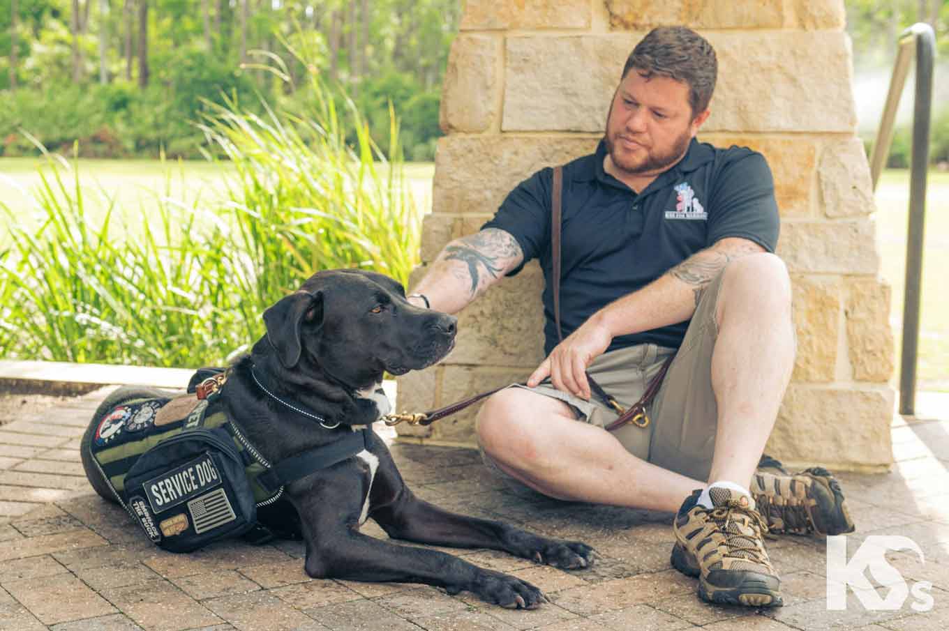 A man sits on the ground with a service dog wearing a vest.